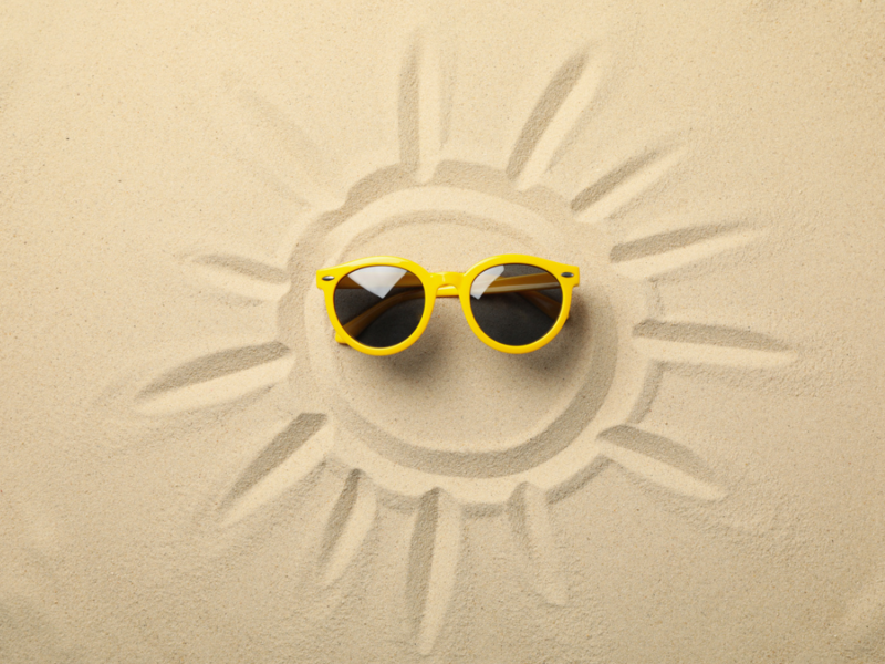 In den feinen Sand wurde ein lachendes Sonnengesicht gemalt. Auf dem gesicht liegt eine gelbe Sonnenbrille. 