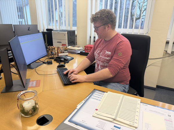 Ein Mann sitzt an einem Schreibtisch und tippt auf einer Tastatur vor zwei Monitoren in einem Büro mit großen Fenstern im Hintergrund.