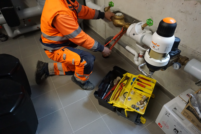 Eine Person in orangefarbener Arbeitskleidung repariert mit einem Rohrschlüssel eine Rohrleitung, während neben ihr ein geöffneter Werkzeugkasten auf dem Boden liegt.