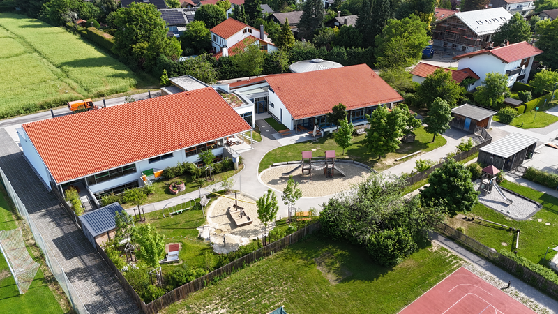 Luftaufnahme eines Schulgeländes mit roten Dächern, Spielplatz, grünen Bereichen und umliegenden Wohnhäusern.