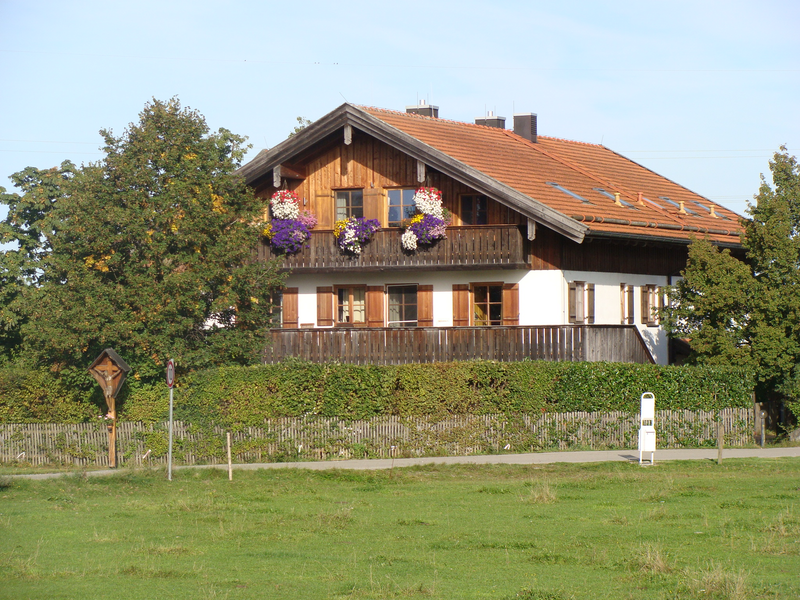 Umgeben von Bäumen und Hecken steht ein großes Haus mit Fensterläden und bepflanzten Balkonen. 