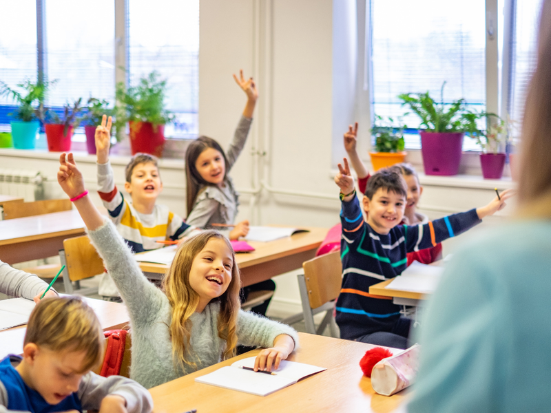 Grundschulkinder sitzen an ihren Tischen in der Klasse und zeigen auf. 