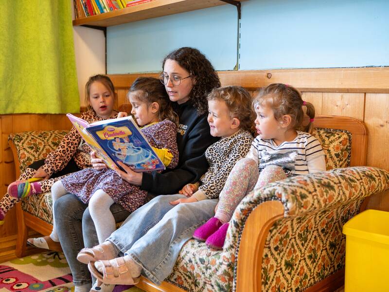 Auf einem großen Sofa sitzen vier Kinder mit einer Erzieherin. Alle schauen in ein Buch. 