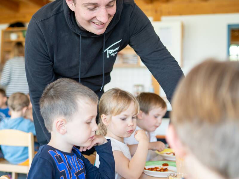 Kleine Kinder sitzen zum Essen am Tisch, ein Erzieher wendet sich einem Kind zu. 