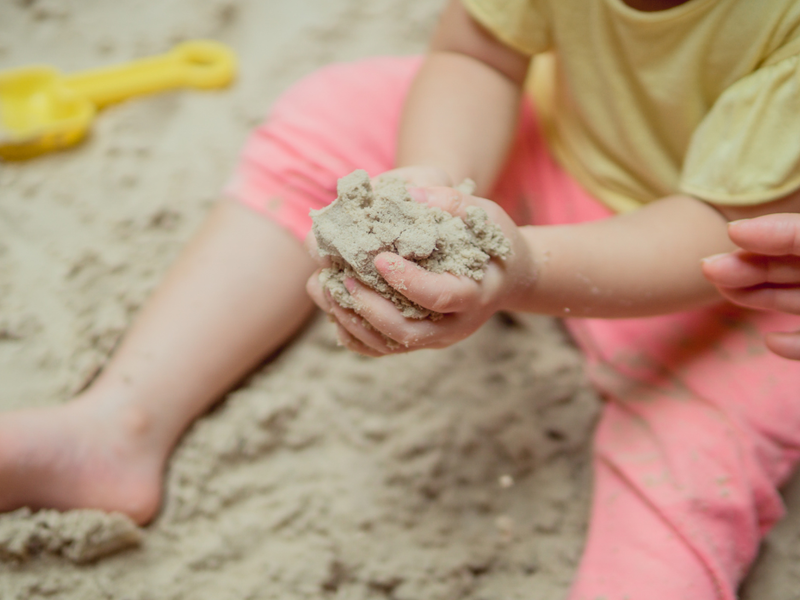Ein kleines Kind sitzt im Sandkasten und hat die Hände voller Sand.