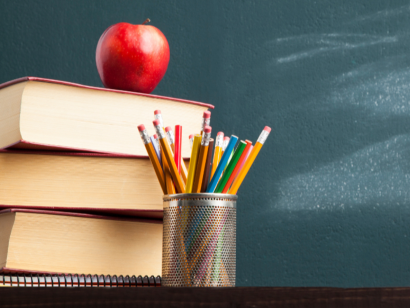 Vor einer Schultafel liegt ein Stapel Bücher, Stifte und ein Apfel. 