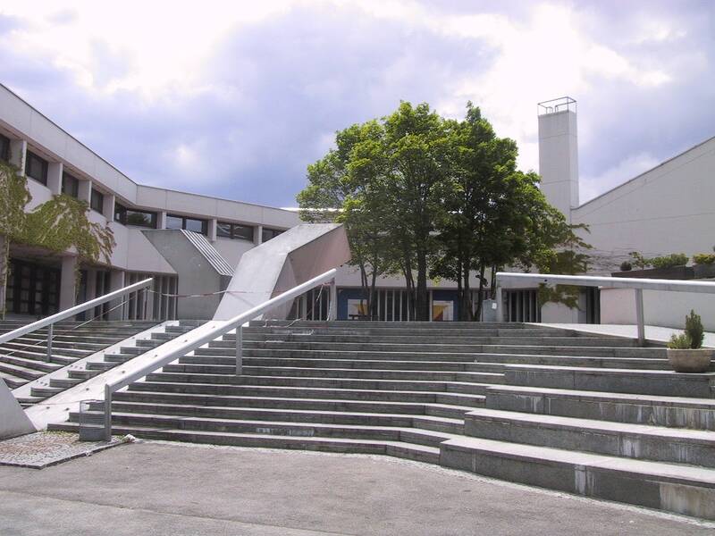 Eine breite Treppe führt zu einem modernen Schulgebäude. Auf dem Schulhof stehen Bäume. 
