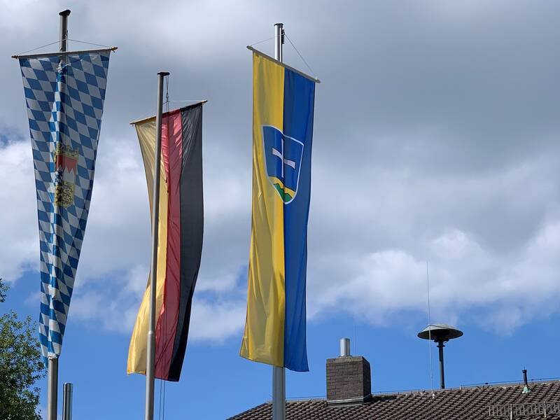 Die Fahne der Bundesrepublik Deutschland, die Fahne des Freistaates Bayern und die Fahne der Gemeinde Straßlach-Dingharting wehen vor einem blauen Himmel.