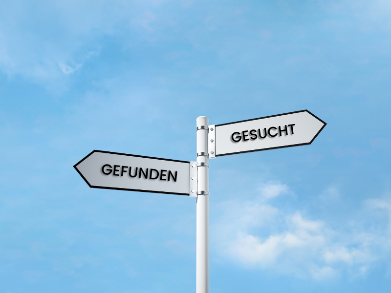 Ein Schild mit zwei Pfeilen zeigt in entgegengesetzte Richtungen, einer mit der Aufschrift "GESUCHT" und der andere mit "GEFUNDEN", vor einem blauen Himmel mit Wolken.