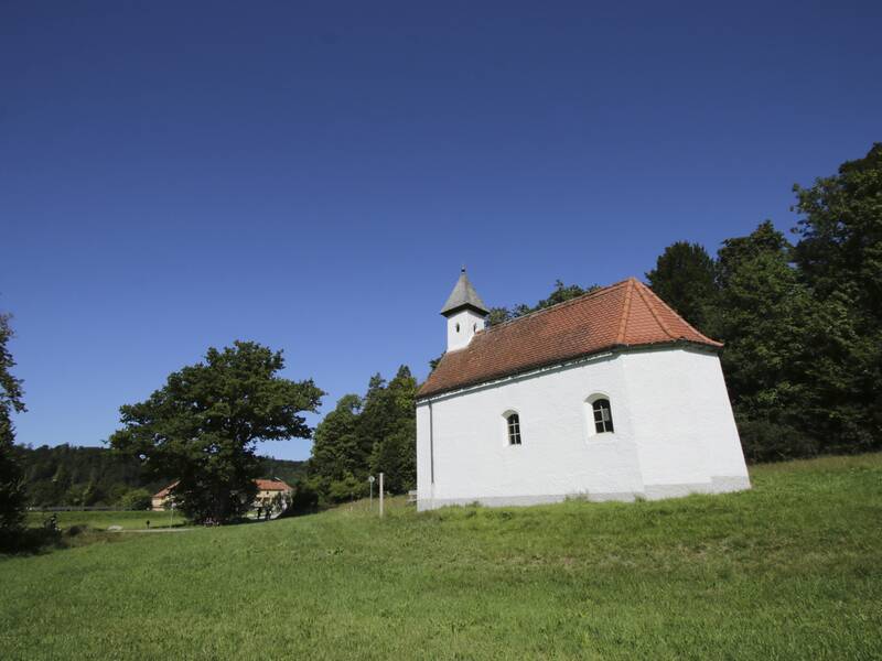 Eine kleine weiße Kapelle steht an einem Waldrand auf einer Wiese.
