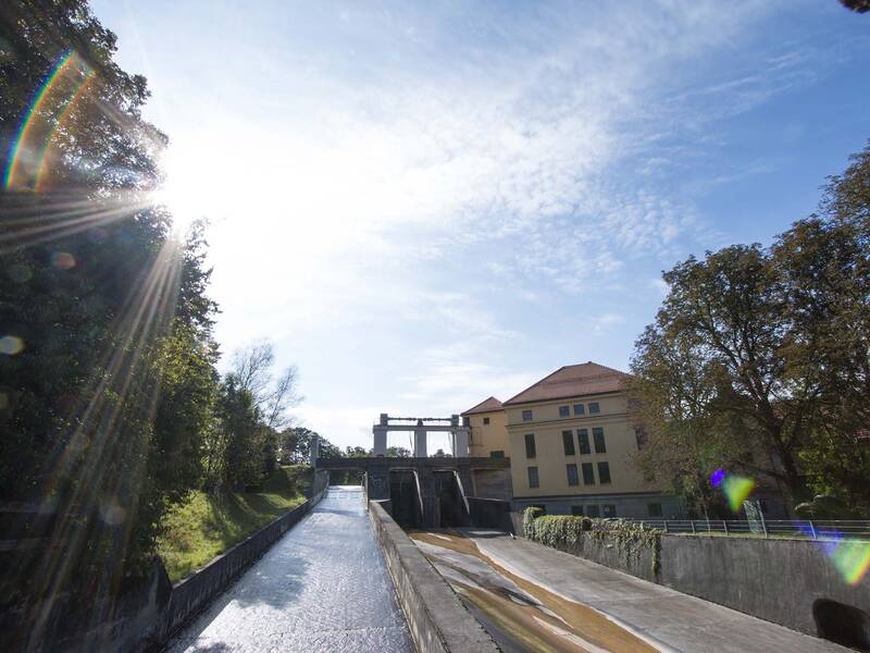 Neben einem gelben Gebäude fließt Wasser eine steile Rampe hinunter. Hier überwinden die Isarfloße den Höhenunterschied am Wasserkraftwerk des Isarkanals-