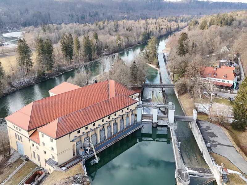 Das Bild zeigt ein weitläufiges Gebäude mit rotem Dach, das an einem Fluss liegt, umgeben von bewaldeter Landschaft.