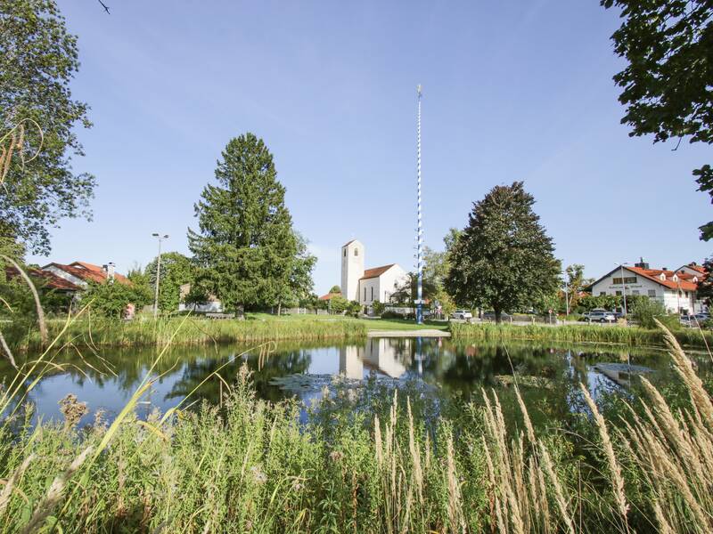 Über einen kleinen Teich hinweg öffnet sich der Blick auf ein Dorf mit Maibaum und Kirche. 
