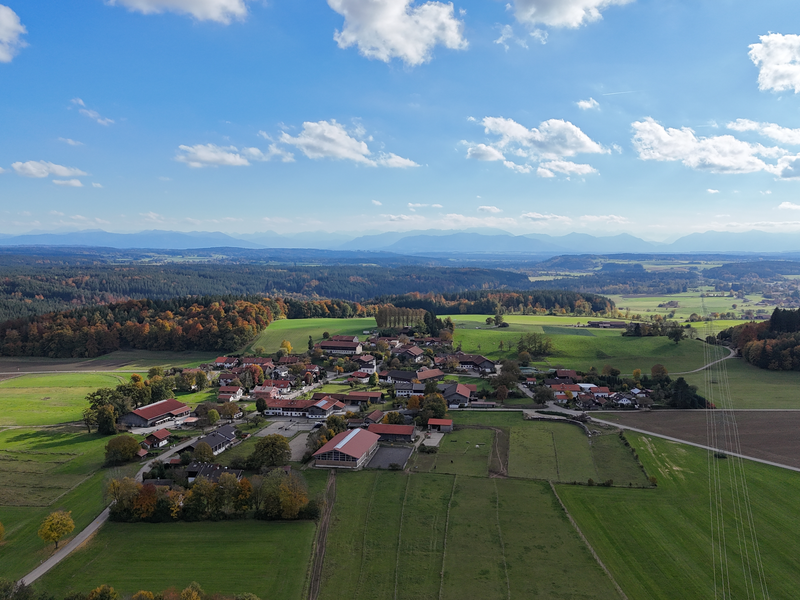 Im Vordergrund ein Teil eines kleinen Ortsteiles mit einer Kapelle. Ringsherum stehen einzelne Baumreihen. Zwischen drinnen Felder in Grün und Braun. 