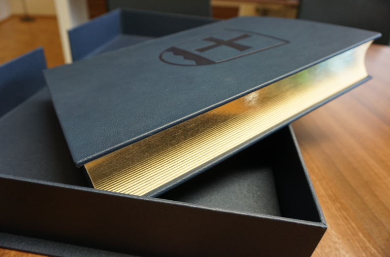 Ein großes Buch mit goldenen Seitenkanten liegt leicht geöffnet in einer passenden Schachtel auf einem Holztisch.