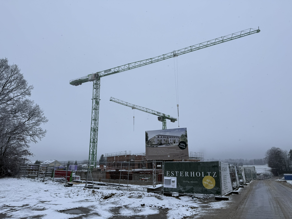 Auf einer verschneiten Baustelle stehen zwei große Kräne und Bauzäune mit Werbebannern, während im Hintergrund ein Gebäude errichtet wird.