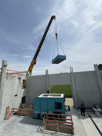 Auf einer Baustelle hebt ein großer Kran ein Bauteil in eine Betonhalle ohne Dach. 