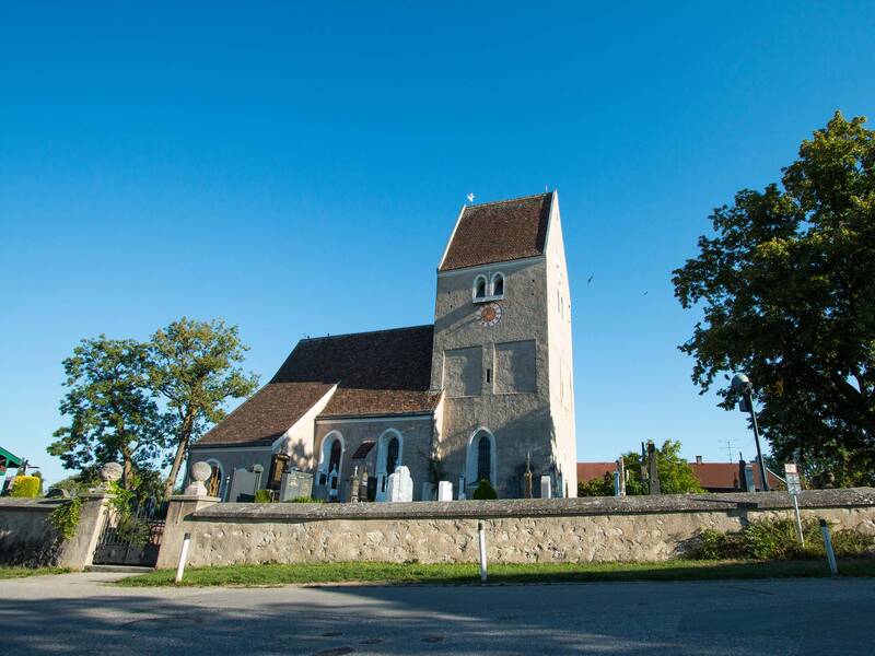 Eine historische Kirche mit einem großen Kirchturm steht vor einem klaren blauen Himmel, umgeben von Bäumen und einer niedrigen Mauer.