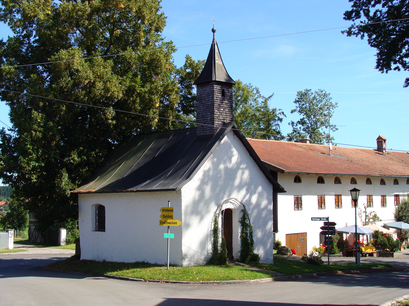 Eine kleine weiße Kapelle mit einem spitzen Turm steht neben einem größeren Gebäude mit roten Dachziegeln, umgeben von Bäumen und einem Wegweiser.