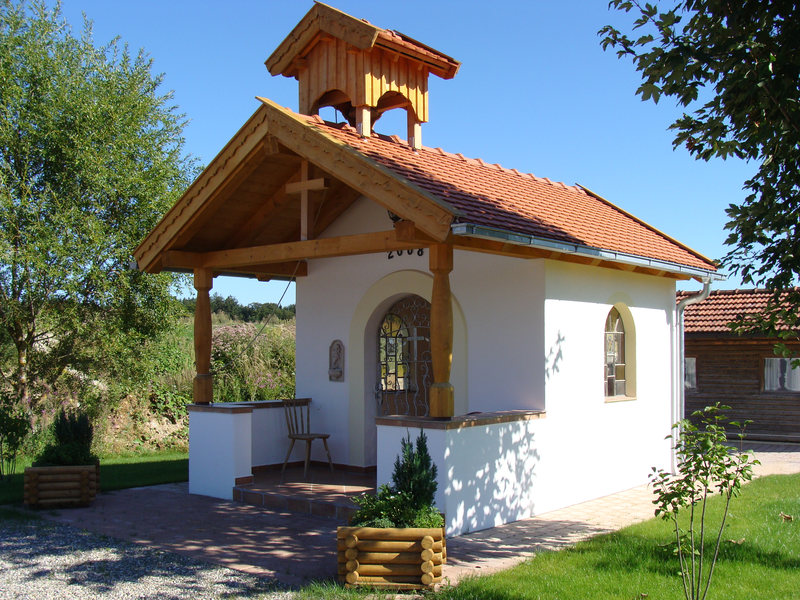 Ein kleines, weißes Kapellengebäude mit einem roten Ziegeldach und einem kleinen Glockenturm steht inmitten grüner Landschaft unter blauem Himmel.