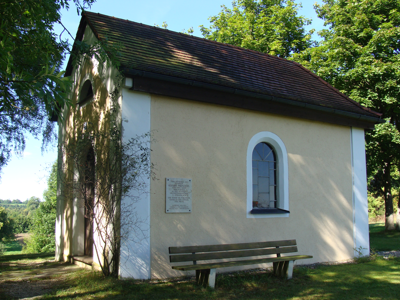 Eine kleine, beige Kapelle mit schrägem Ziegeldach steht in einer grünen Landschaft, umgeben von Bäumen, mit einer Bank an der Seite und einem runden Fenster.