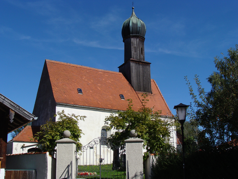 Eine Kirche mit einem roten Ziegeldach und einem charakteristischen Turm steht vor einem klaren blauen Himmel, umgeben von Bäumen und einem schmiedeeisernen Tor.