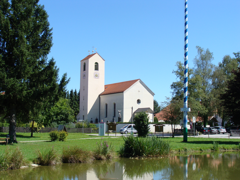 Vor einem klaren blauen Himmel steht eine weiße Kirche mit rotem Ziegeldach, umgeben von Bäumen und einem kleinen Teich im Vordergrund.