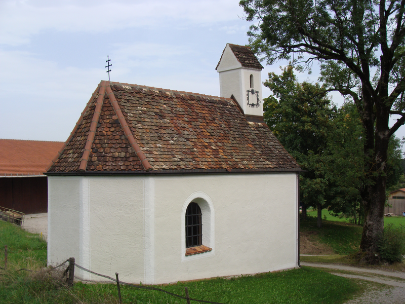 Eine kleine, weiß verputzte Kapelle mit einem ziegelgedeckten Dach und Glockenturm steht in einer ländlichen Umgebung neben einem Baum.