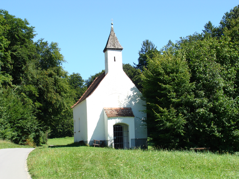 Eine kleine weiße Kapelle mit einem spitzen Dach steht am Waldrand, umgeben von grünen Bäumen unter einem klaren, blauen Himmel.