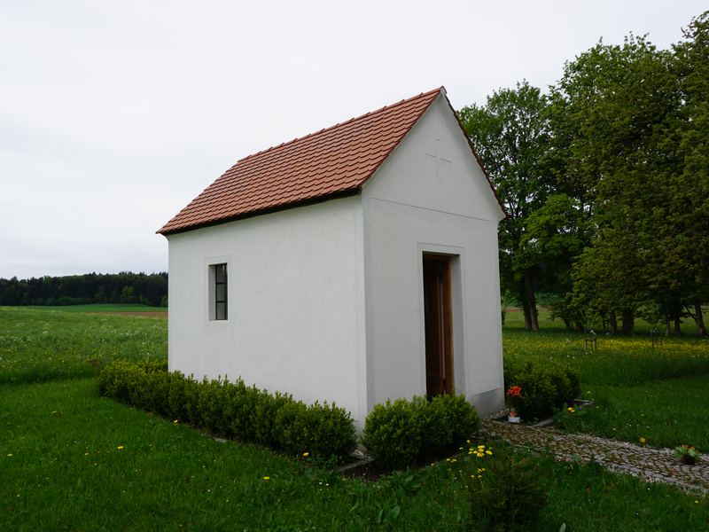 Eine kleine, weiße Kapelle mit rotem Ziegeldach steht inmitten einer grünen Wiese, umgeben von Bäumen.
