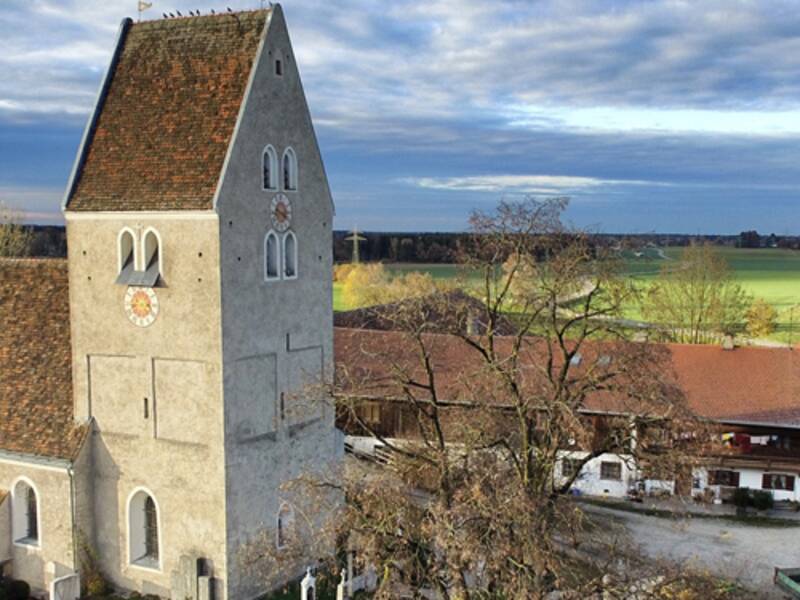Luftaufnahme einer romanischen Kirche mit wehrhaftem Turm am Rande eines Dorfes umgeben von Wiesen. 