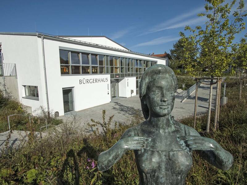 Eine Skulptur einer Frau steht im Vordergrund, während im Hintergrund ein Gebäude mit der Aufschrift "Bürgerhaus" von Bäumen umgeben sichtbar ist.