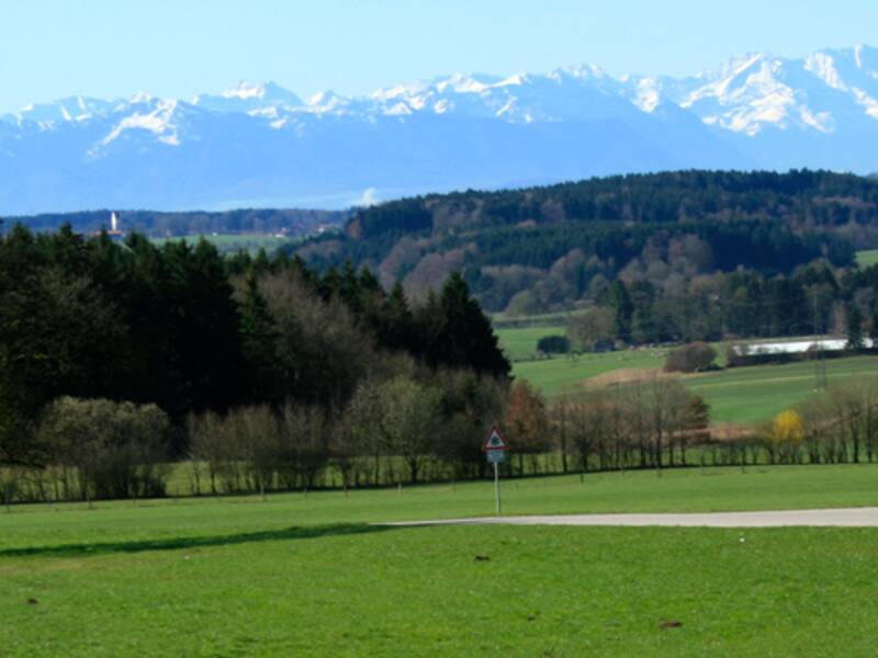Eine sanft geschwungene Landstraße führt durch grüne Wiesen und Wälder, mit einem Dorf im Hintergrund und einer imposanten Bergkette am Horizont.