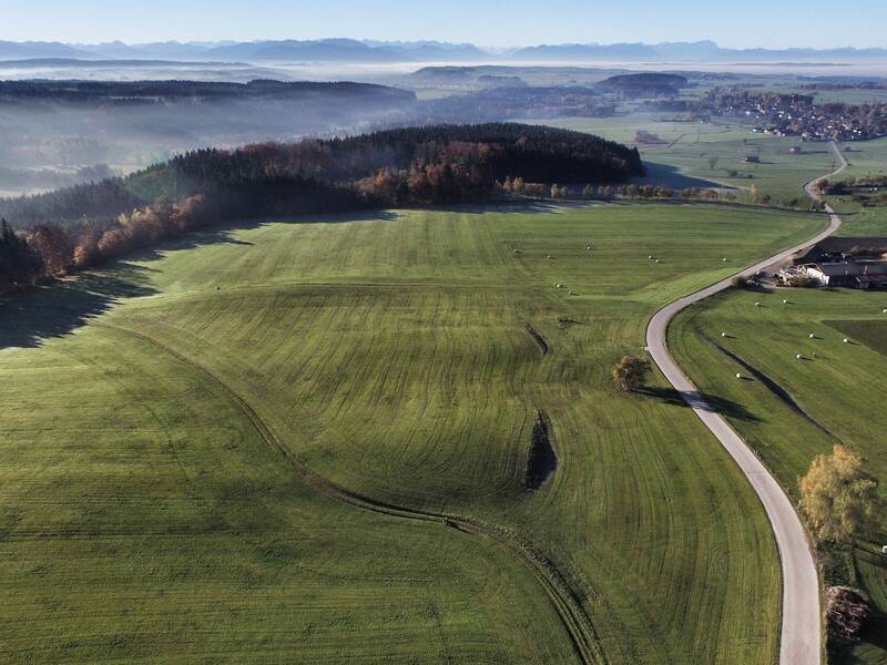 Das Bild zeigt eine weitläufige, grüne Landschaft mit Feldern, Wäldern, einem schlängelnden Weg und einem kleinen Dorf im Hintergrund unter einem klaren Himmel.