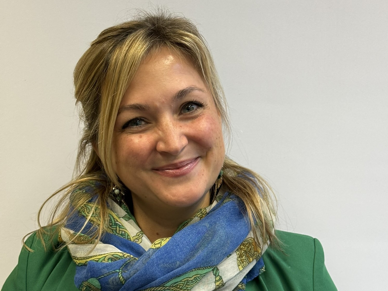 	  Eine lächelnde Person mit blondem Haar trägt einen grünen Blazer und einen blau-weiß gemusterten Schal vor einem neutralen Hintergrund.