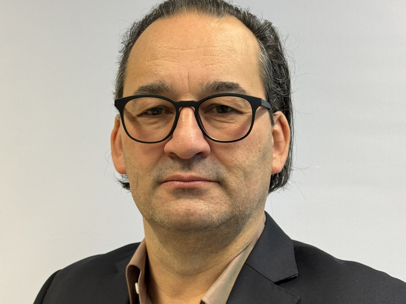 Ein Mann mit Brille und dunklem, zurückgekämmtem Haar trägt einen schwarzen Blazer vor einem neutralen Hintergrund.