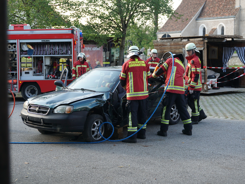 Fünf Feuerwehrmänner vor einem havarierten Auto beim demonstrieren einer Hilfsaktion. Ein Feuerwehrauto steht links im Hintergrund